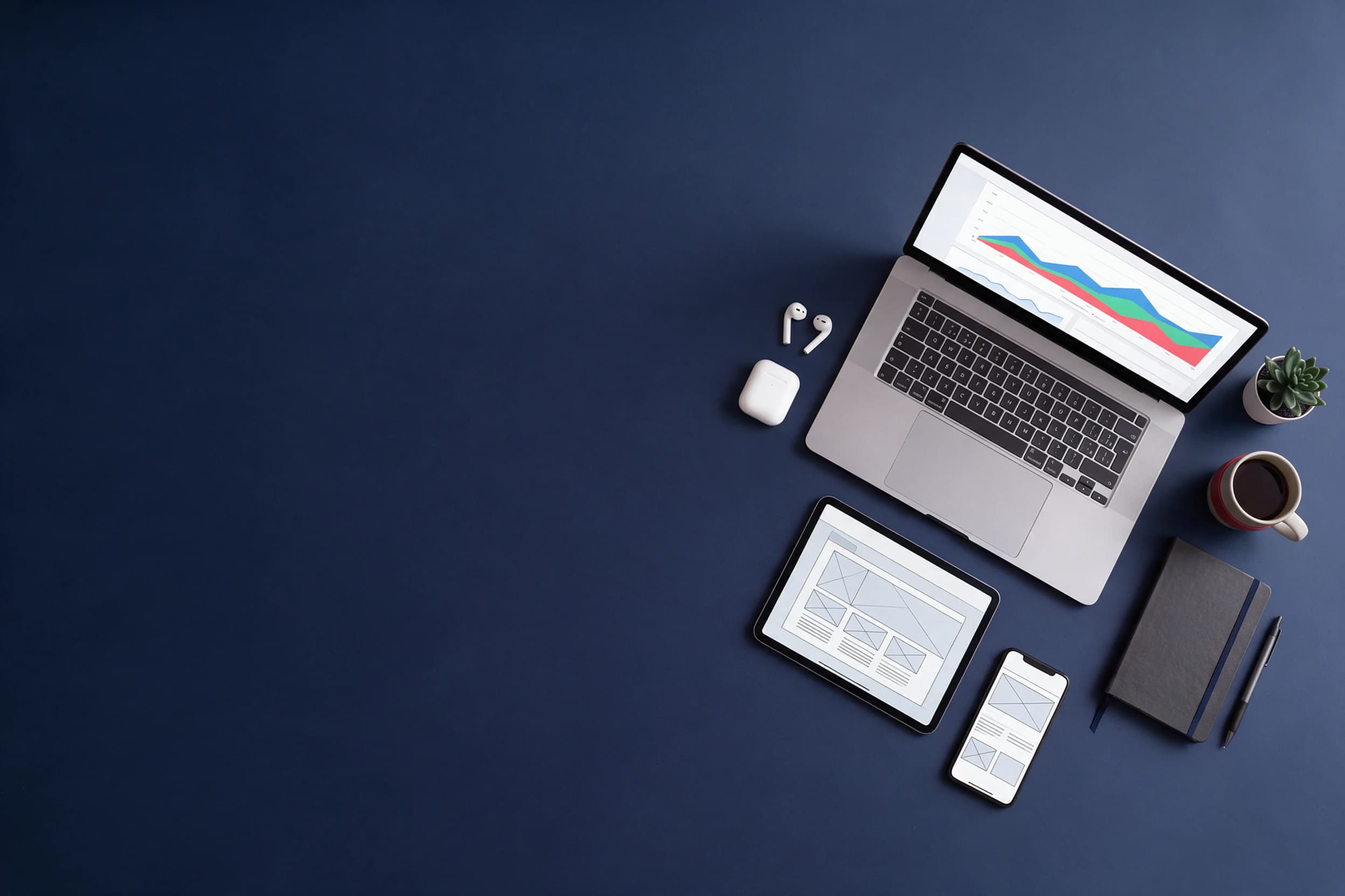 Overhead view of a laptop with an analytics dashboard, a tablet with a website layout and a phone with a social feed on a navy desk